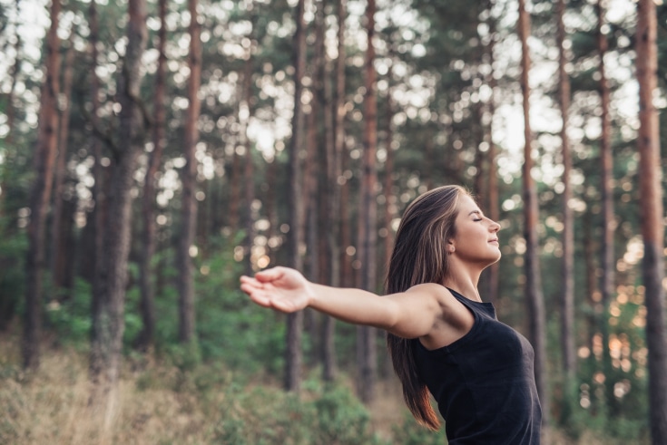 Young woman raising her arms to nature Attractive young brunette woman standing among trees in a forest with her eyes closed and arms raised up to embrace nature